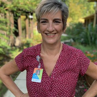 A woman with short hair, wearing a polka dot shirt and an ID badge, smiles while standing in a garden.