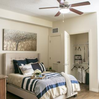 A cozy bedroom with a neatly made bed, stylish linens, and a ceiling fan. The room features a small closet and wall artwork.