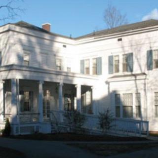 A historic white house with large windows and a porch, surrounded by trees.