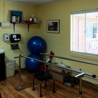 A well-equipped physical therapy room featuring parallel bars, a blue exercise ball, and ample natural light.