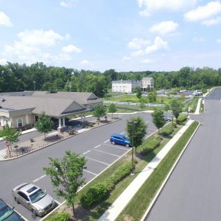 Aerial view of a parking lot with multiple cars and buildings surrounded by trees under a partly cloudy sky.