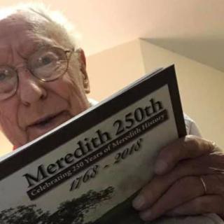 An elderly person wearing glasses is reading a booklet titled 'Meredith 250th'.