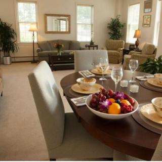 A modern living and dining area with a round table set with plates, glasses, and a fruit bowl, surrounded by comfortable chairs.