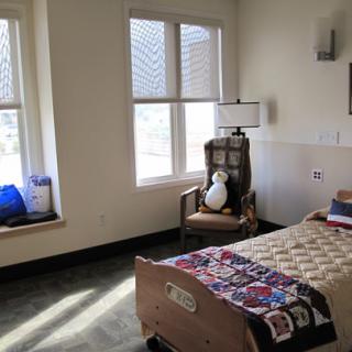 A hospital room with a bed, quilt, chair, and large windows.