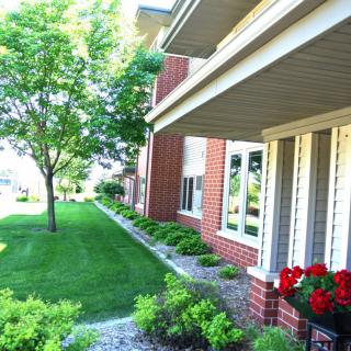 A well-maintained yard with green lawn, trees, and a flower bed in front of a brick house.