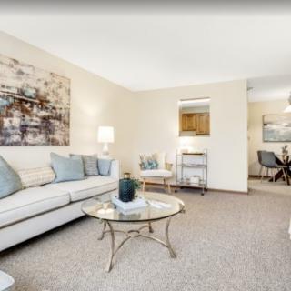 A cozy living room with neutral tones, featuring a sofa and glass coffee table, accented by artwork and soft lighting.