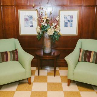 A cozy seating area featuring two green chairs with striped cushions, a wooden side table with a floral arrangement, and framed artwork on the wooden paneled wall.