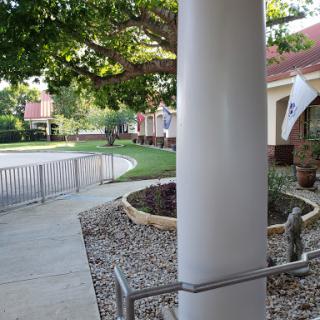 A view of a pathway leading to a building, framed by a column and surrounded by greenery.