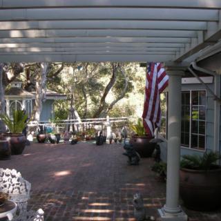 A serene outdoor patio area with a pergola, brick flooring, and various potted plants under dappled sunlight.