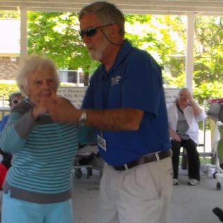 An elderly couple dancing outdoors, enjoying a sunny day.