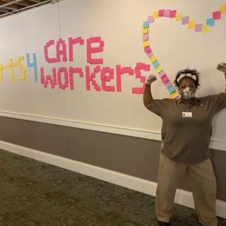 A healthcare worker poses in front of a wall adorned with post-it notes spelling 'care workers' and forming a heart shape.