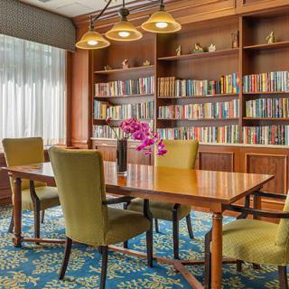 A cozy library reading room with a wooden table, upholstered chairs, and shelves filled with books.