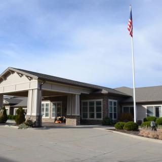 An exterior view of a building with an American flag raised on a flagpole, featuring a landscaped entrance under a clear sky.