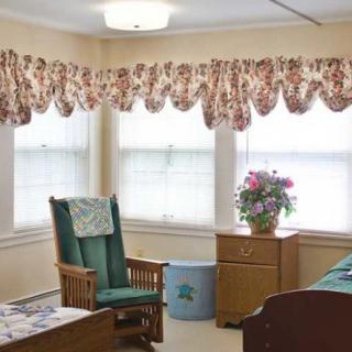 A cozy living room with a green cushioned chair, wooden furniture, floral curtains, and a potted plant on a dresser.
