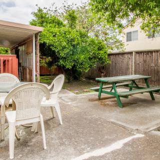 A backyard with a picnic table and plastic chairs on a concrete patio, surrounded by trees and a wooden fence.