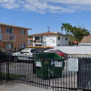 A parking lot in front of apartment buildings with dumpsters and a fence, under a cloudy sky.