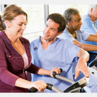 A physical therapist assists a patient on exercise equipment, with other patients exercising in the background.