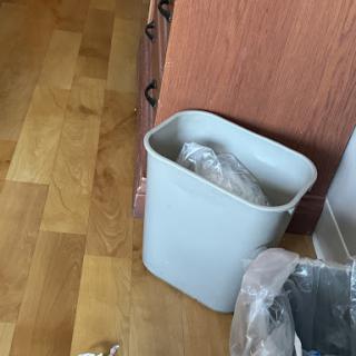 A gray trash can sits on a wooden floor, with some waste visible inside.