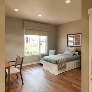 A cozy bedroom with a single bed, desk, and chair, featuring wooden flooring and a large window.