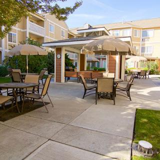 A cozy outdoor patio area with tables and chairs, shaded by umbrellas, set within an apartment complex.