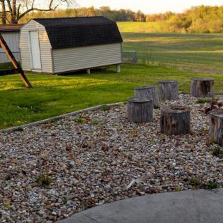 A backyard scene featuring a shed, fire pit area with wooden stumps, and gravel, set against an open field.