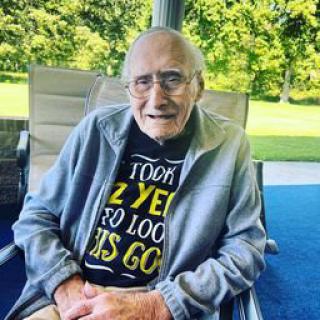 An elderly man sitting outdoors, wearing glasses and a t-shirt with a cheerful message.
