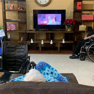 An elderly person wrapped in a blanket watching television in a care facility, with another individual in a wheelchair nearby.
