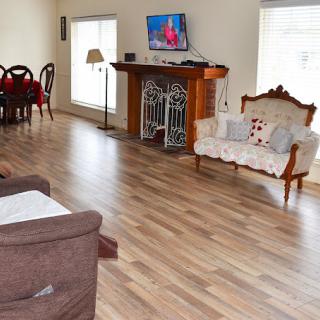 A spacious living room with hardwood floors, featuring a sofa, armchair, dining table, and a television mounted above a fireplace.