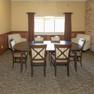 A dining room with a round table and six chairs, featuring a carpeted floor and a window with curtains.