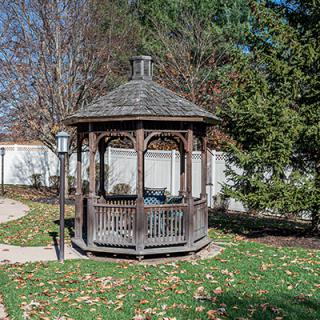 A wooden gazebo surrounded by trees and fallen autumn leaves in a garden setting.