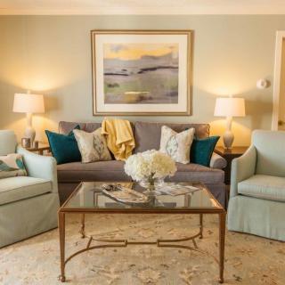 A cozy living room with a sofa and two armchairs, complemented by a glass coffee table, decorative cushions, ambient lighting from lamps, and a painting on the wall.