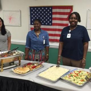 Three people standing behind a buffet table with various dishes, in front of an American flag.