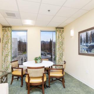 A cozy dining area with a round table surrounded by chairs, adorned with a plant and flanked by large windows and artwork.