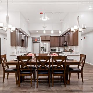 A modern dining area with a spacious kitchen in the background, featuring wooden furniture and elegant chandeliers.