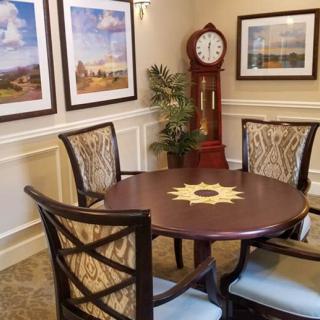 A cozy dining area with a circular wooden table surrounded by four chairs, featuring a grandfather clock and framed wall art.