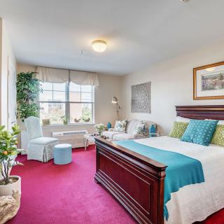 A bright bedroom with a red carpet, featuring a large bed, armchair, and window with curtains.