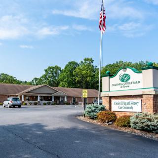 A one-story building with a parking lot and an American flag, surrounded by trees, under a clear blue sky.