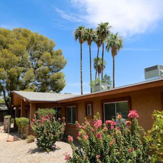 A single-story house surrounded by flowers and palm trees on a sunny day.