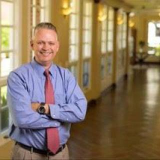 A man in business attire smiling in a well-lit office corridor.