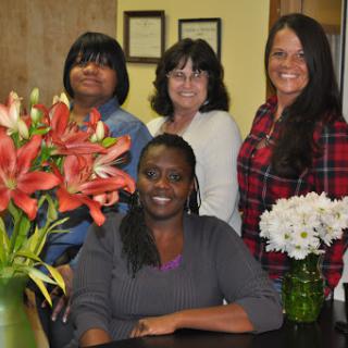 A group of four women posing together in an office setting, surrounded by beautiful flower arrangements.