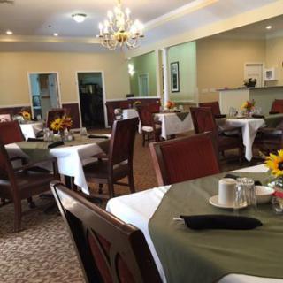A well-decorated dining area featuring multiple tables with white and green tablecloths, surrounded by wooden chairs and adorned with sunflowers under a chandelier.