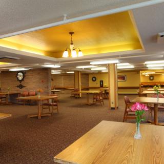 An empty dining hall with wooden tables and chairs, warm lighting, and decorative elements on the walls.