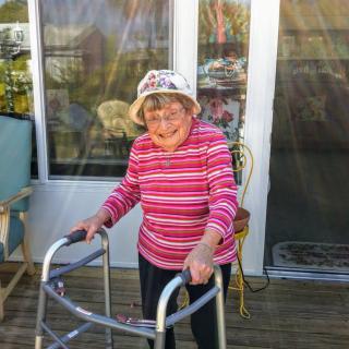 An elderly woman with a walker, wearing a striped shirt and hat, stands on a porch.
