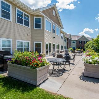 A spacious outdoor patio area with tables, chairs, and planters adjacent to a modern two-story building on a sunny day.