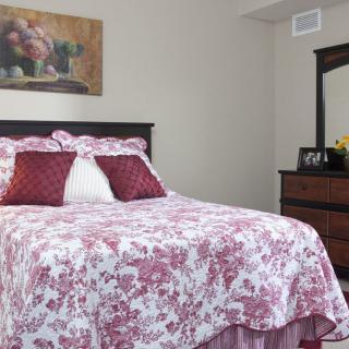 A cozy bedroom featuring a bed with floral bedding and decorative pillows, alongside a wooden dresser and mirror.