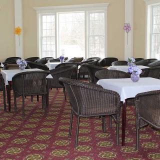 A well-decorated dining room with tables and chairs arranged neatly, featuring large windows and carpeted flooring.