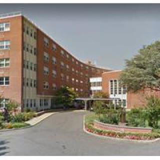 The image shows the exterior of a hospital building with an entrance and landscaped surroundings.