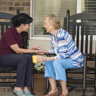A caregiver and an elderly woman share a conversation while seated on rocking chairs outside.