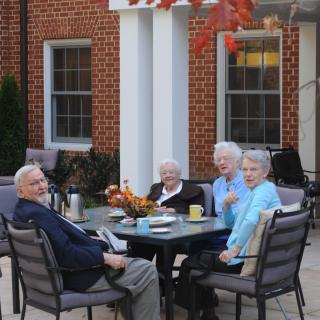A group of seniors enjoying a social gathering outdoors during autumn, sitting at a table near a brick building.