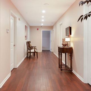 A well-lit hallway with wooden flooring, featuring a chair and a small table with a lamp.